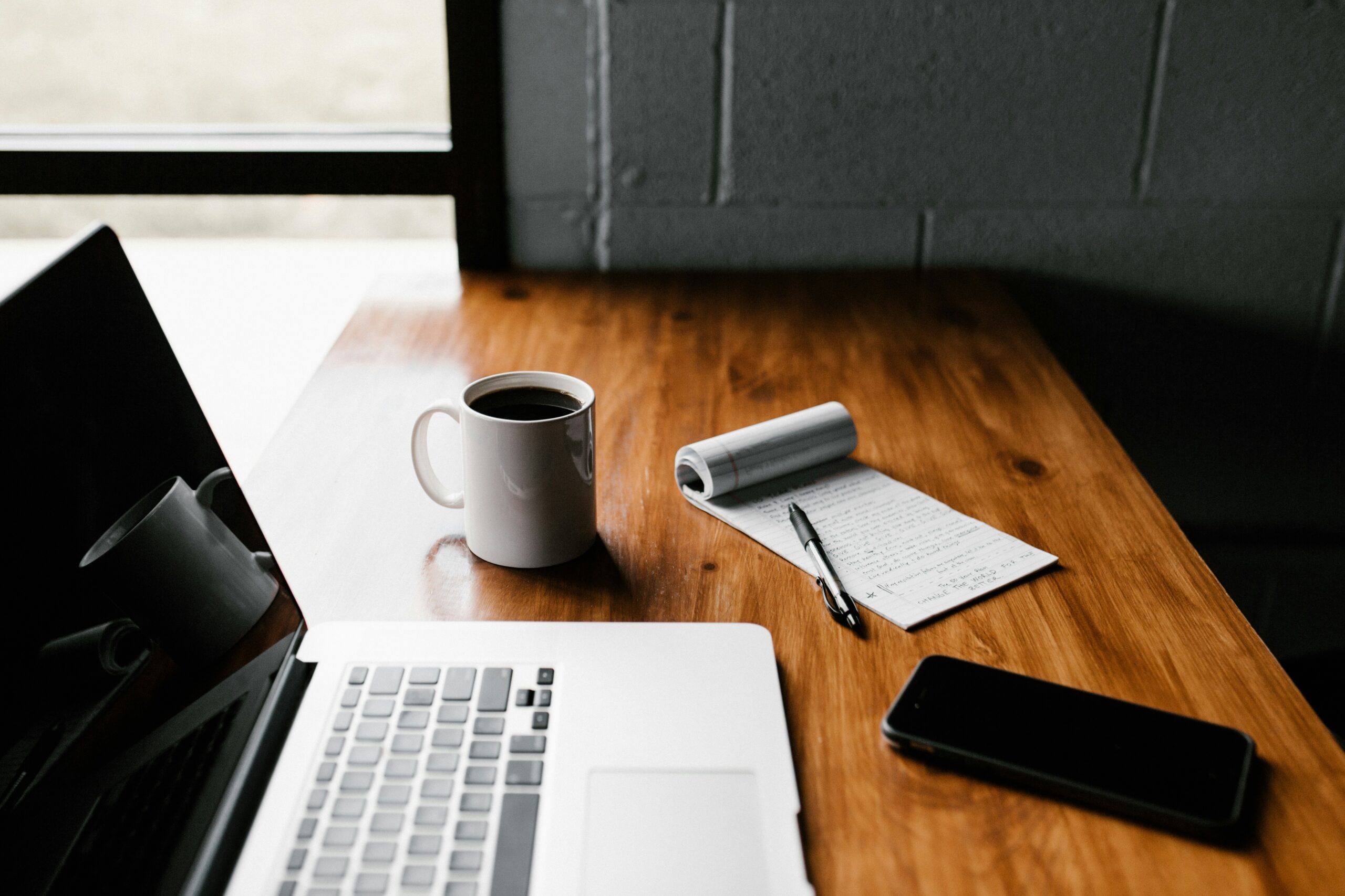 Minimalist workspace with laptop, coffee mug, notepad, pen, and smartphone on a wooden desk near a window