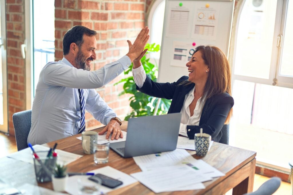 Business colleagues high-fiving in a modern office after successful meeting, with laptop and documents on table