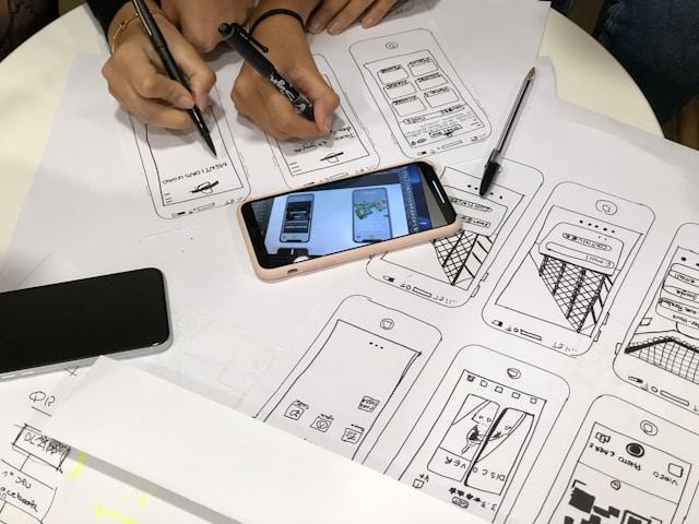 Two hands sketching mobile app designs on various pieces of paper. 